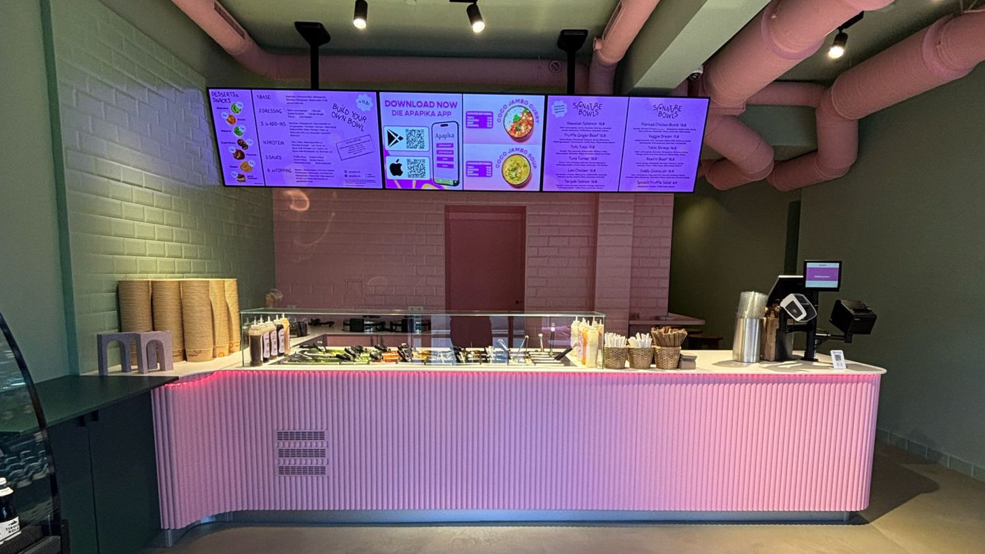 Restaurant mit modernem pink beleuchtetem Tresen, Salatbar und digitalen Menüs auf Bildschirmen unter rosa Deckenrohren.