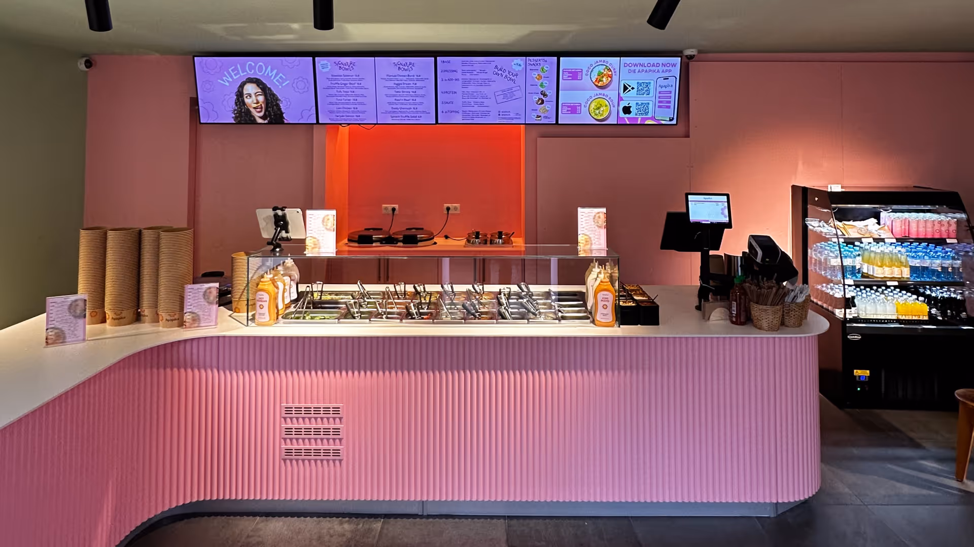 Modernes pinkfarbenes Restaurant mit Salad-Bar, digitale Menüs an Wand und Getränkekühlschrank.