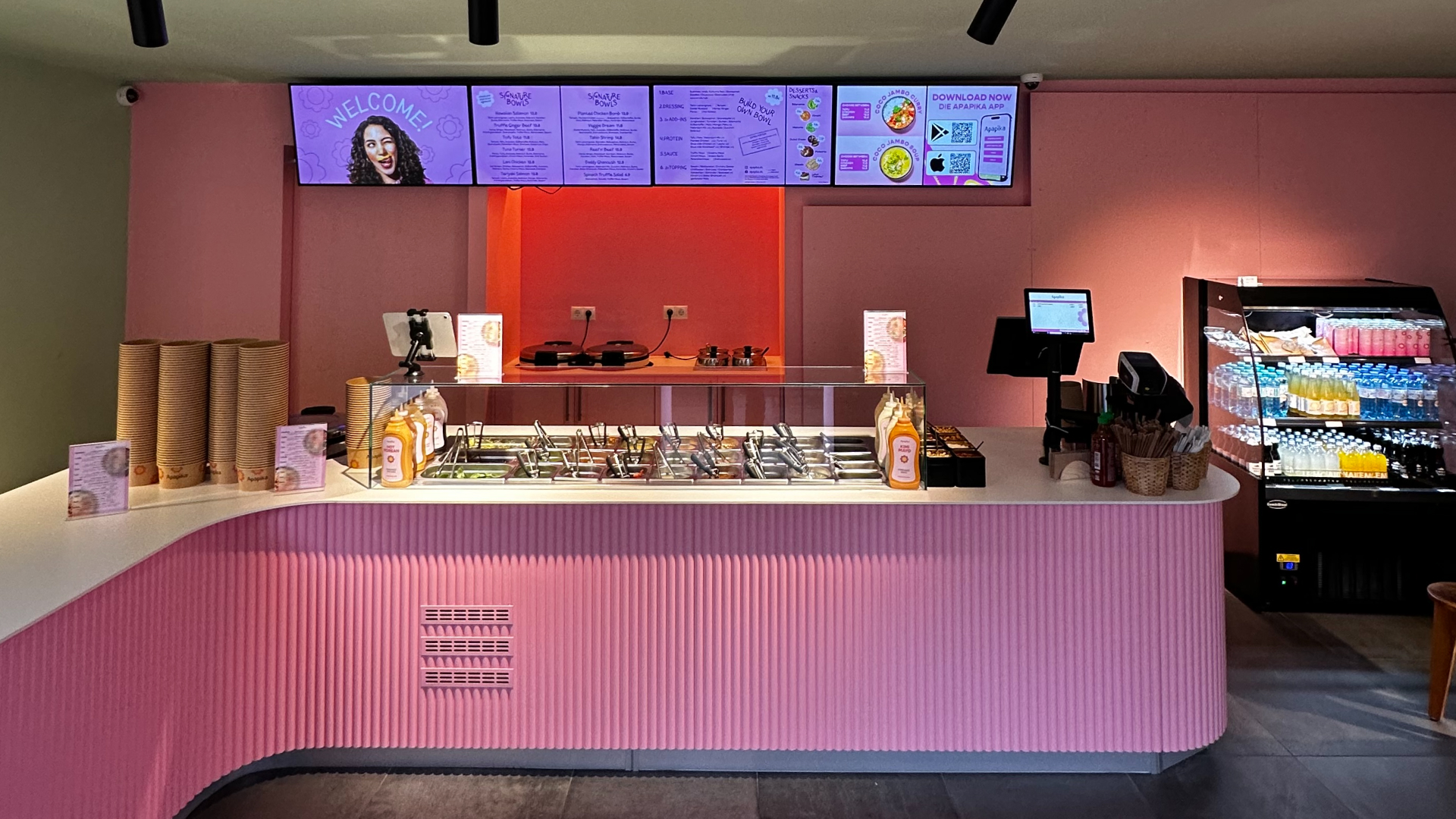 Modernes pinkfarbenes Restaurant mit Salad-Bar, digitale Menüs an Wand und Getränkekühlschrank.