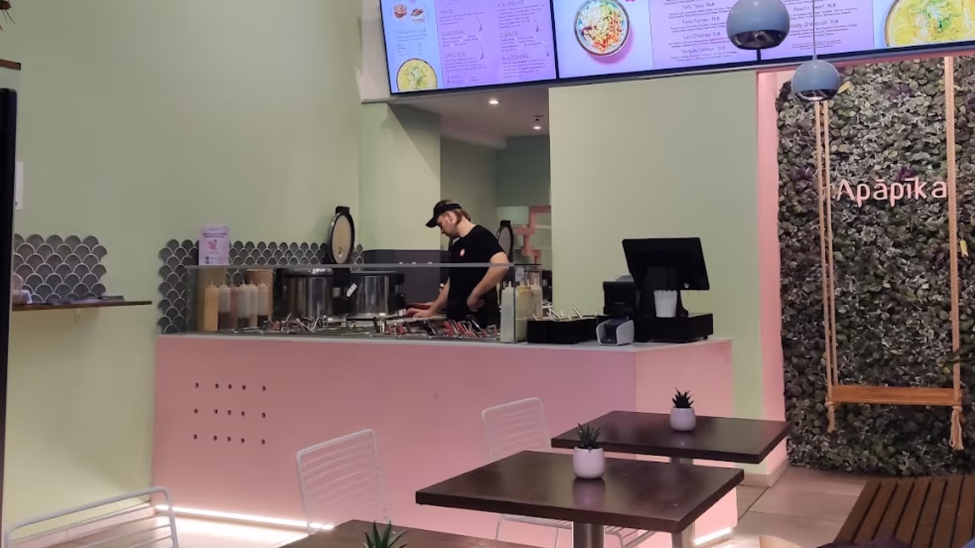 Moderne Restauranttheke mit einem Mitarbeiter, der hinter der Glasabdeckung verschiedene Speisen zubereitet, und Sitzbereich mit Tischen und Pflanzen.