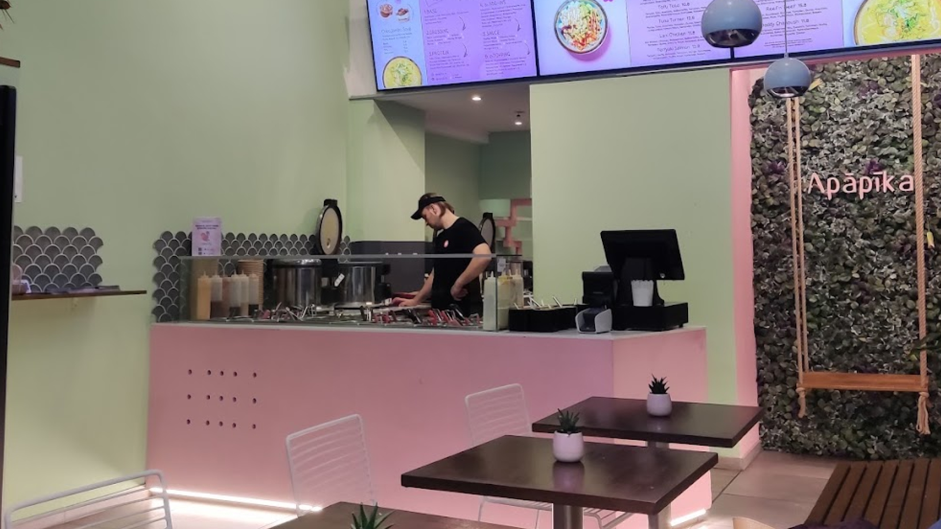 Moderne Restauranttheke mit einem Mitarbeiter, der hinter der Glasabdeckung verschiedene Speisen zubereitet, und Sitzbereich mit Tischen und Pflanzen.