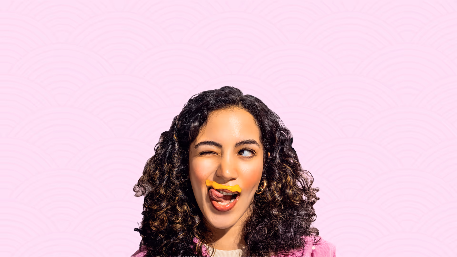 Frau mit lockigem Haar und pinkem Mantel zwinkert, mit einem Klecks Senf über der Oberlippe.