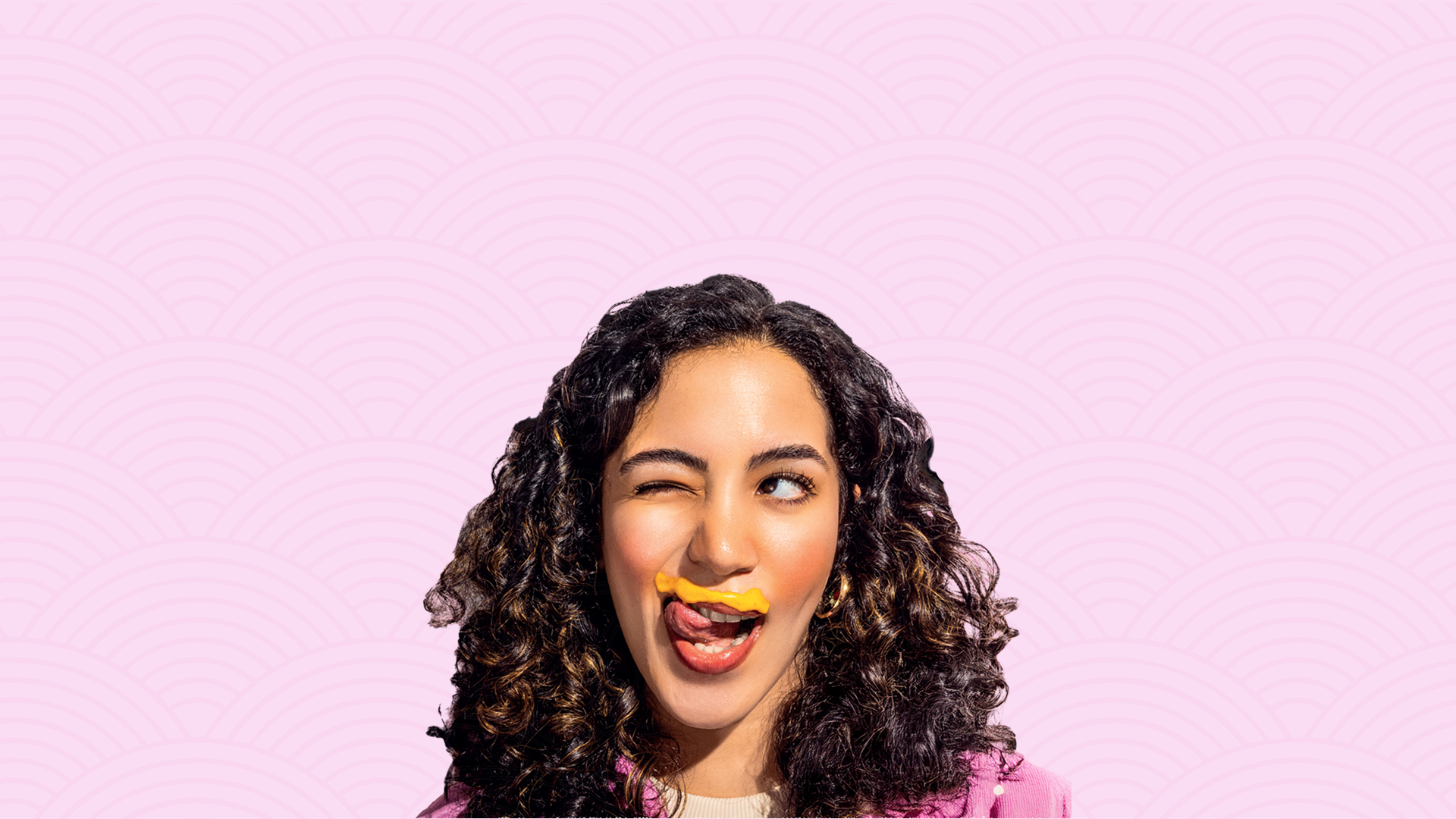 Frau mit lockigem Haar und pinkem Mantel zwinkert, mit einem Klecks Senf über der Oberlippe.