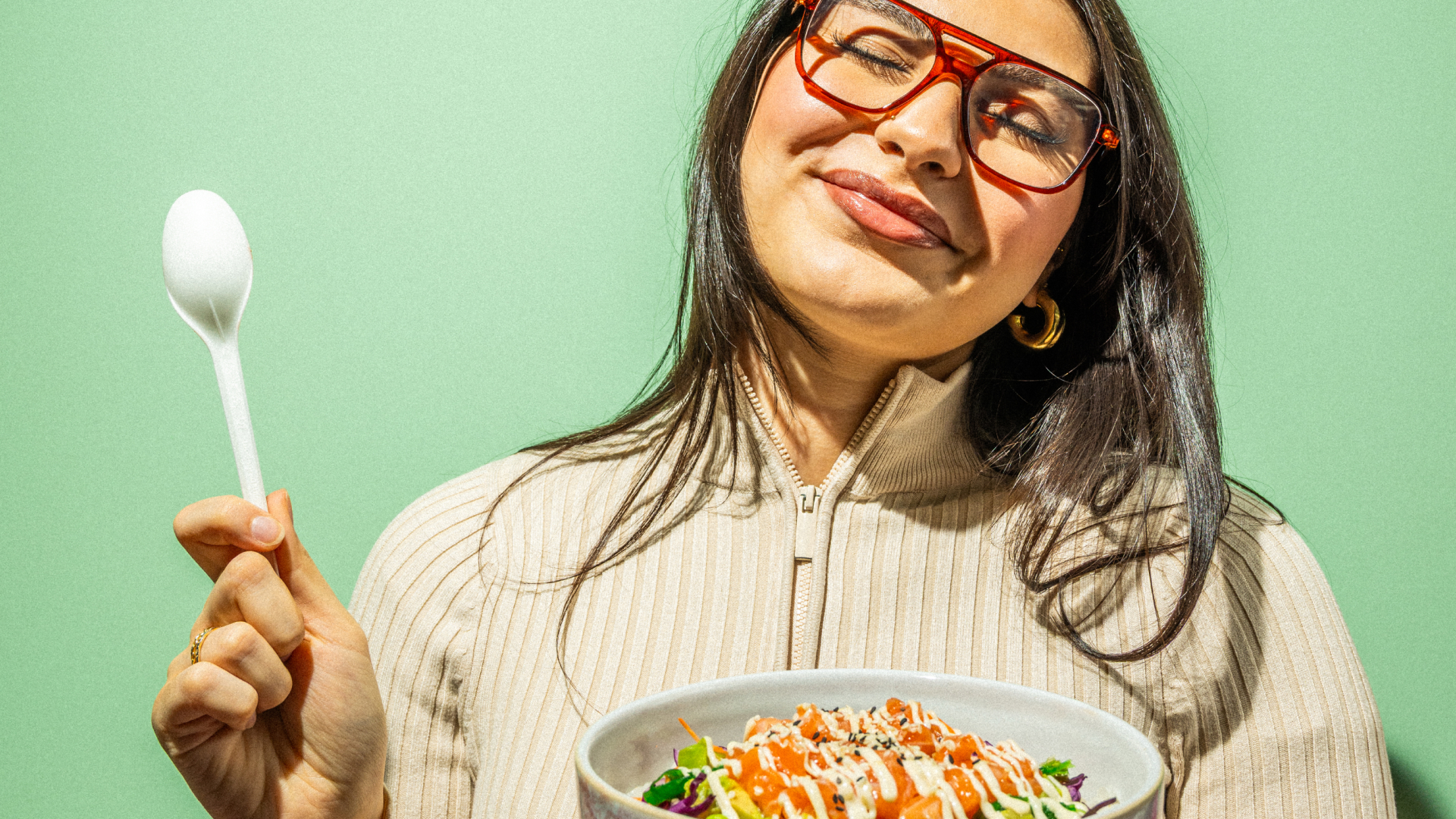 Frau mit roten Brillen hält einen weißen Löffel und eine Schüssel mit frischem, bunt garniertem Salat vor grünem Hintergrund.
