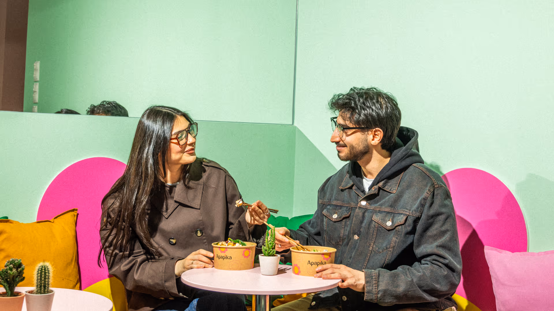 Ein Mann und eine Frau mit Brille essen gemeinsam mit Stäbchen aus Apapika-Schalen an einem kleinen runden Tisch mit Kakteen in einem hellgrünen Raum.