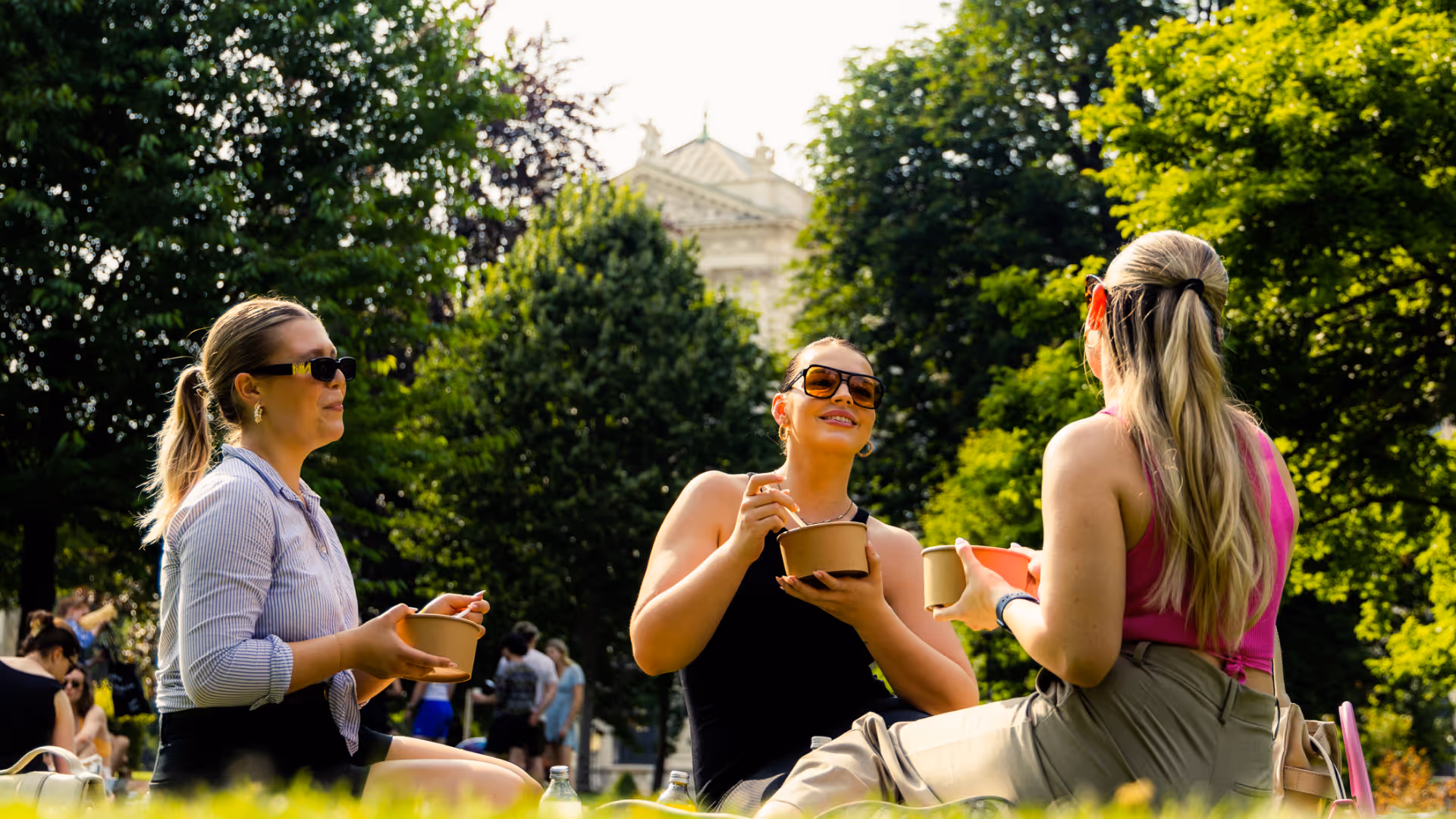 Drei Frauen sitzen im Park auf der Wiese, essen aus Schalen und unterhalten sich bei sonnigem Wetter.