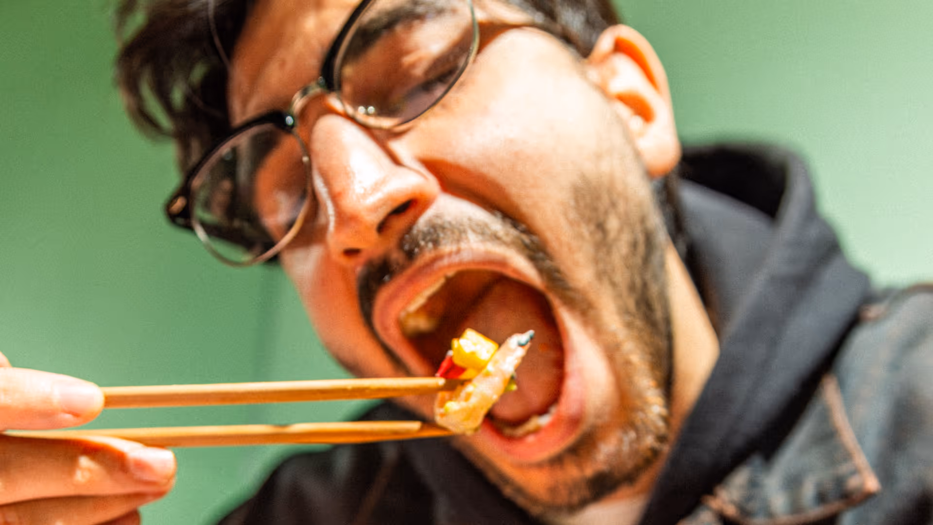 Nahaufnahme eines Mannes mit Brille, der mit Chopsticks ein Stück Sushi isst.