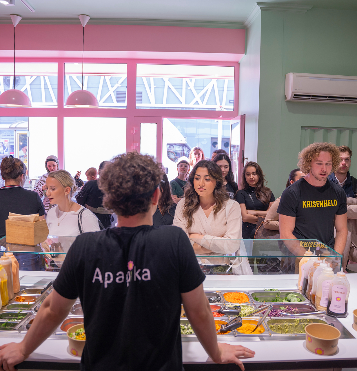 Menschen stehen in einem hellen Restaurant vor einer Theke mit frischen Zutaten und Saucen, bedienen von einem Mitarbeiter mit Apapika-Shirt.