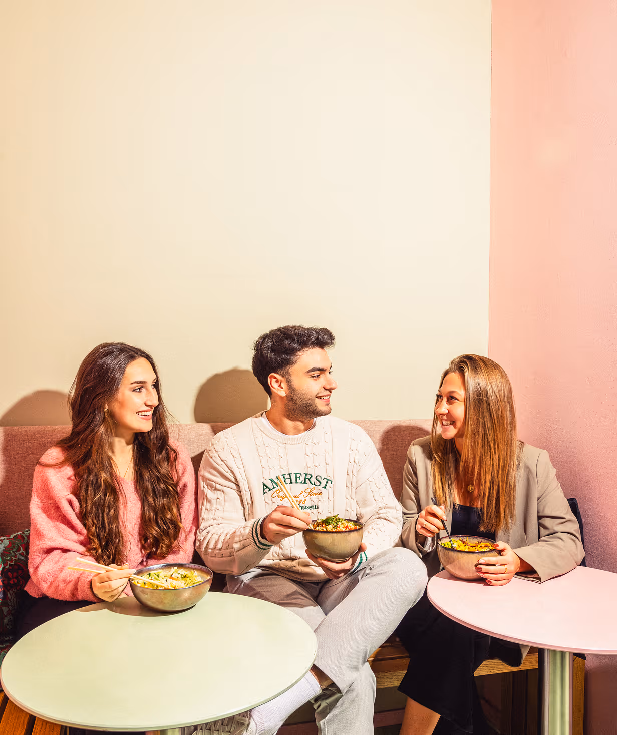 Drei junge Menschen sitzen zusammen in einem Café und essen Gerichte aus Schalen, während sie sich unterhalten und lächeln.