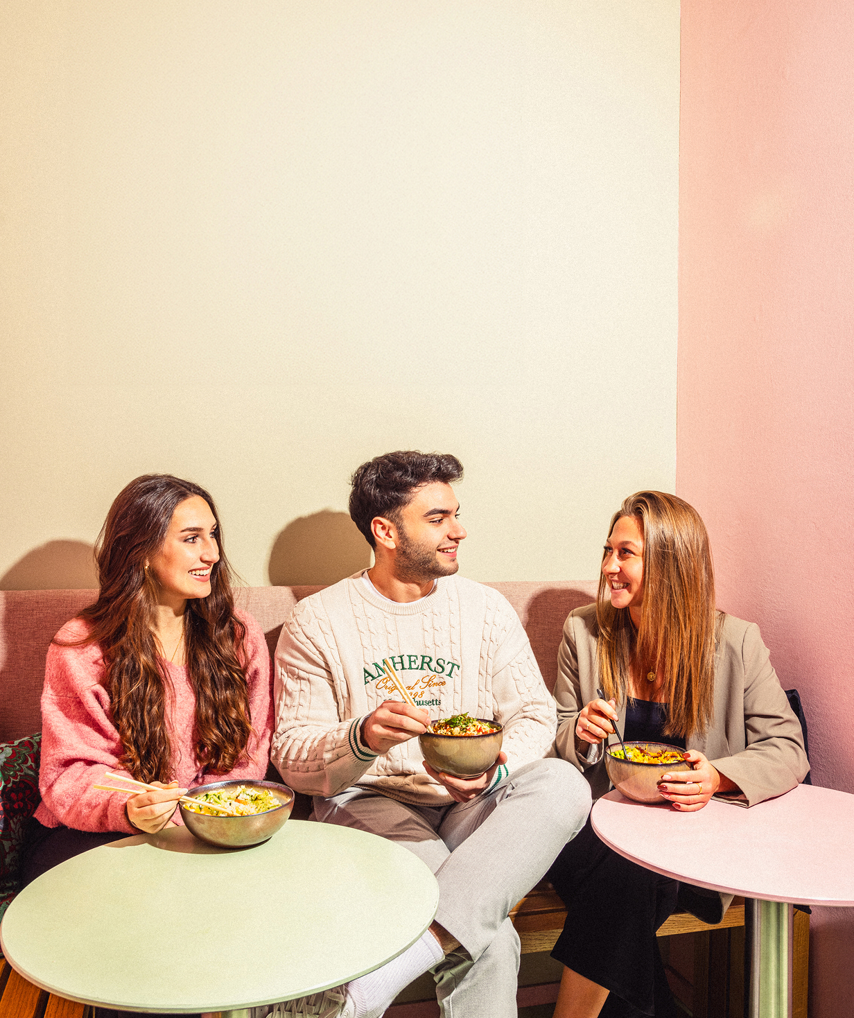Drei junge Menschen sitzen zusammen in einem Café und essen Gerichte aus Schalen, während sie sich unterhalten und lächeln.