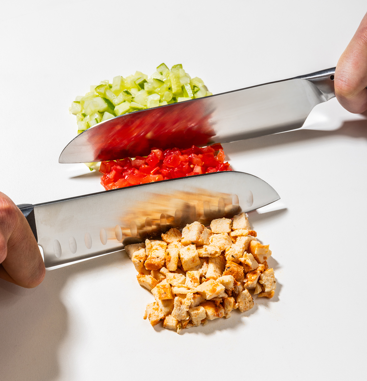 Zwei Hände schneiden mit großen Messern fein gewürfeltes Hühnerfleisch, Tomaten und Gurken auf einem weißen Schneidebrett.