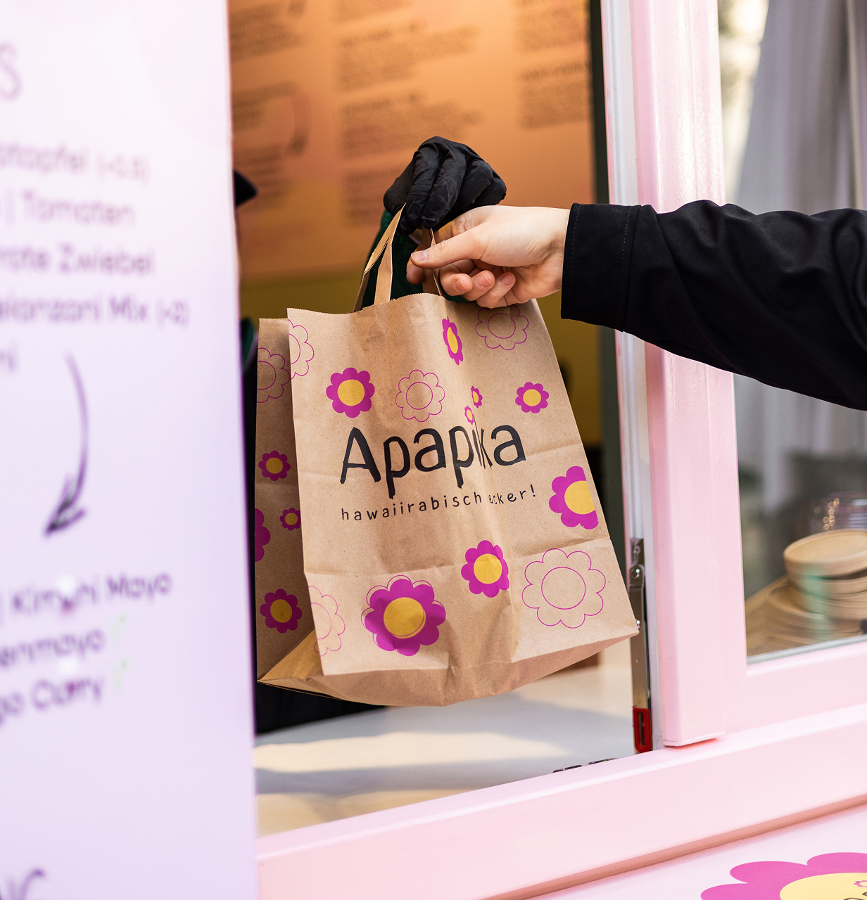 Eine Hand in schwarzer Kleidung erhält eine Papiertüte mit dem Aufdruck 'Apapia' und Blumen von einer Person mit schwarzem Handschuh an einem pinken Fenster.