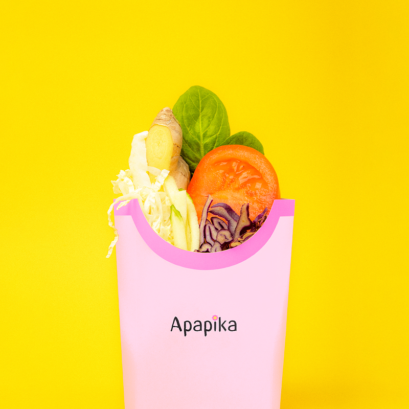 Bunte frische Gemüseblätter, Ingwer und Tomatenscheibe in einer rosa Verpackung mit Aufschrift Apapika vor gelbem Hintergrund.
