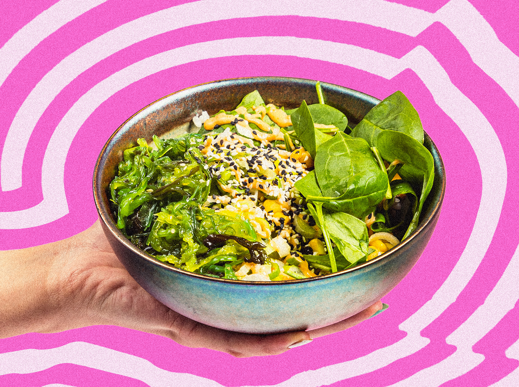 Hand hält eine Schüssel mit frischem Salat, Algen, Sesamsamen und Spinatblättern vor einem pink-weißen Wellendesign-Hintergrund.