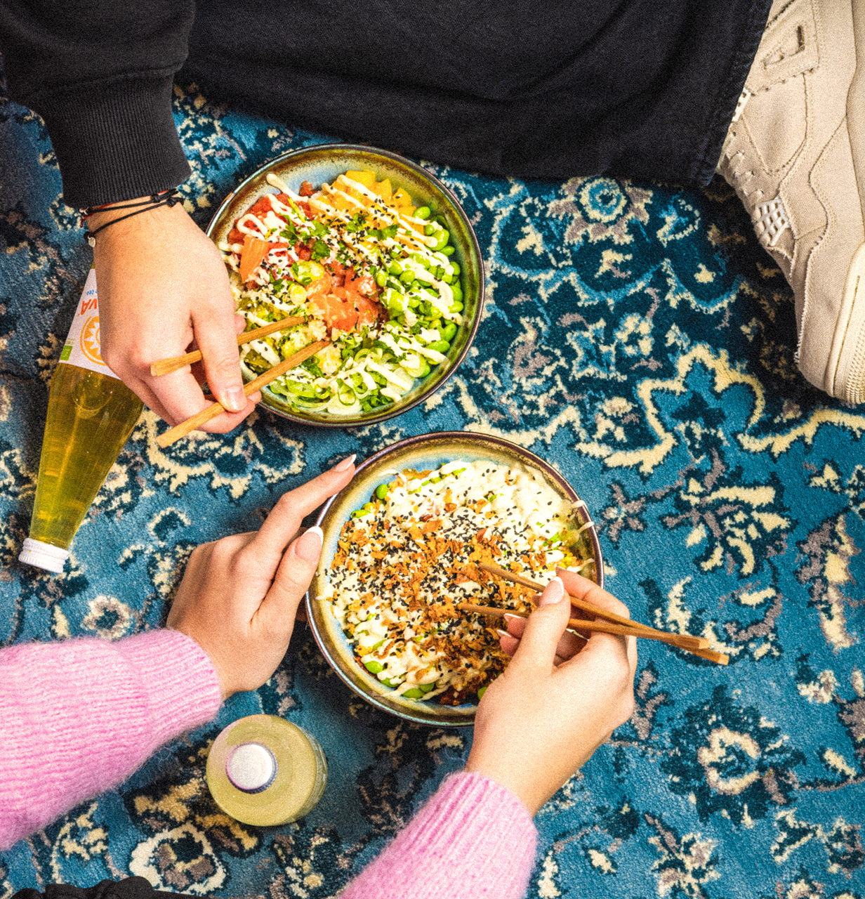 Zwei Personen sitzen auf einem blau gemusterten Teppich und essen bunte Poké-Bowls mit Essstäbchen, daneben stehen zwei Flaschen Getränke.
