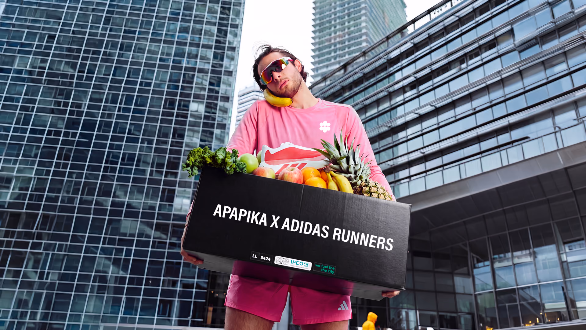 Mann in pinker Sportkleidung hält eine Kiste mit Obst und Gemüse vor Hochhäusern.