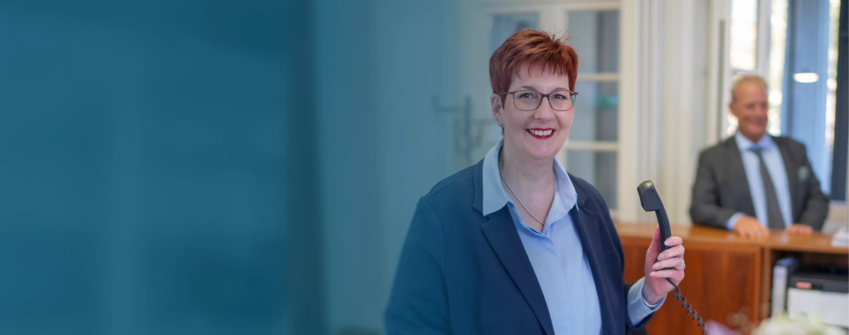 Lächelnde Frau mit rotem kurzen Haar und Brille hält Telefonhörer im Empfangsbereich der GPP Treuhand