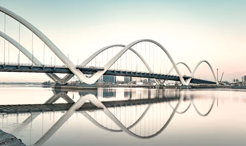 Un pont moderne avec des arches blanches se reflète dans une eau calme au coucher du soleil.