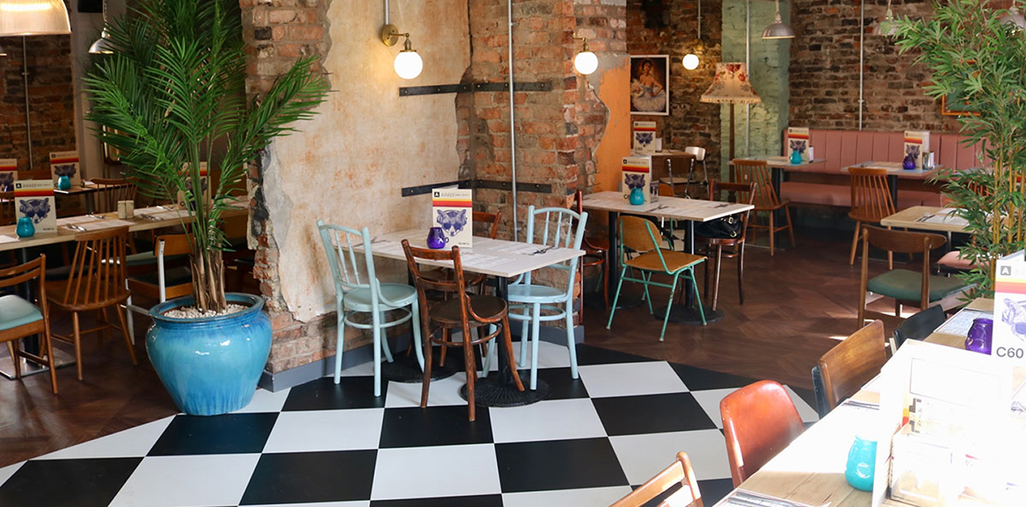 Cozy restaurant interior with exposed brick walls, mixed wooden and painted chairs, checkered black and white floor, and potted plants.