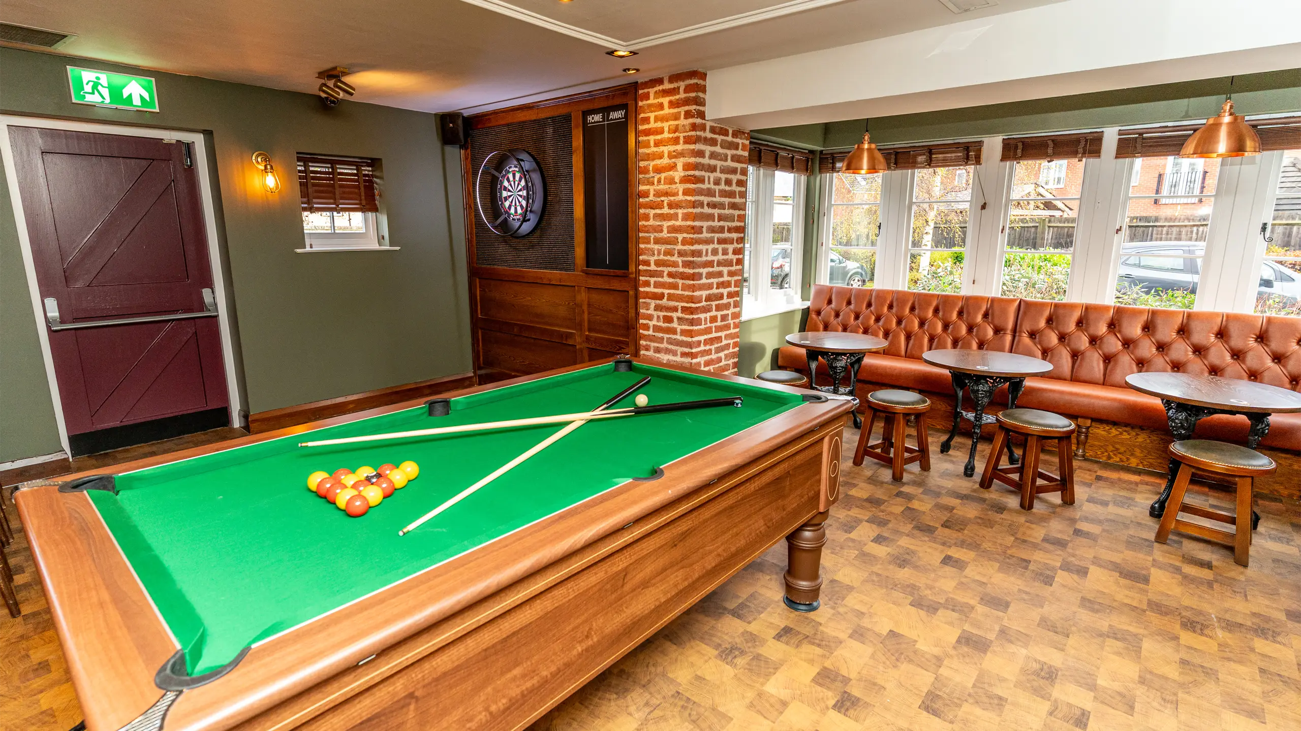 Indoor game room with a green felt pool table, two crossed cues, a dartboard on the wall, and leather seating with wooden stools by windows.