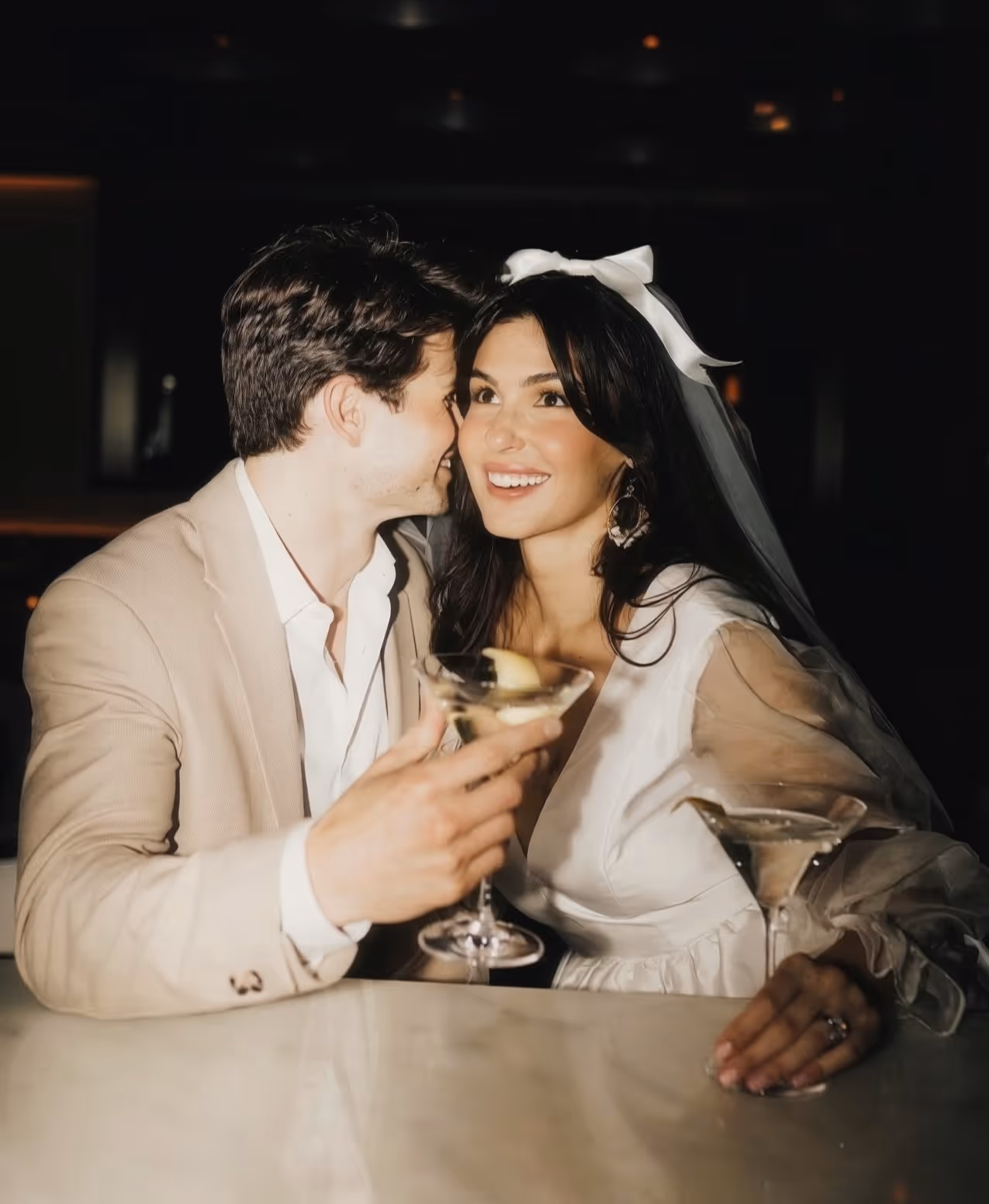 Couple at a bar with cocktails, the woman dressed in a white bridal outfit with veil and bow, smiling brightly.