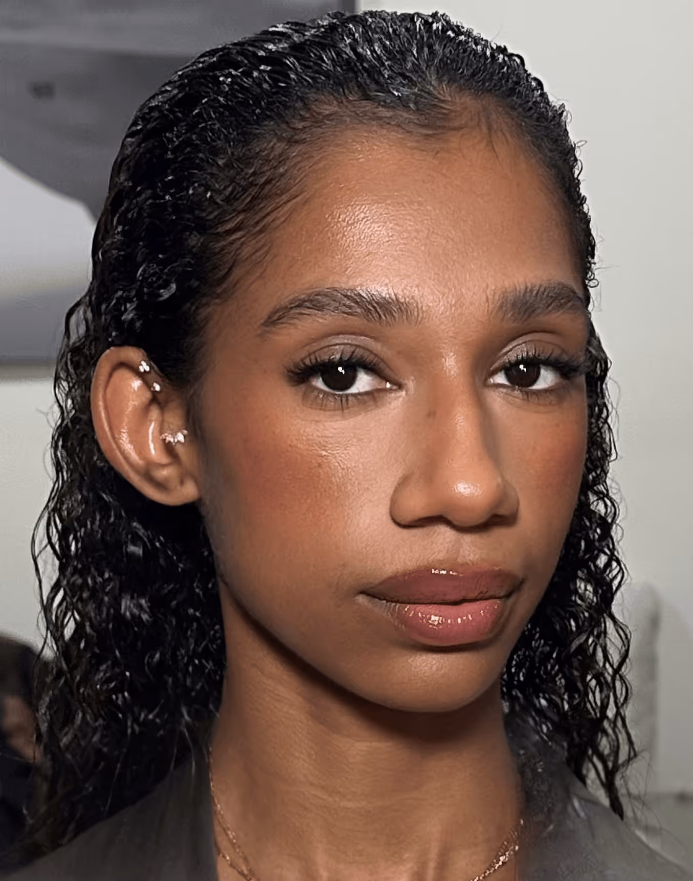 Close-up of a young woman with dark curly wet hair, natural makeup, pink lip gloss, and multiple ear piercings.