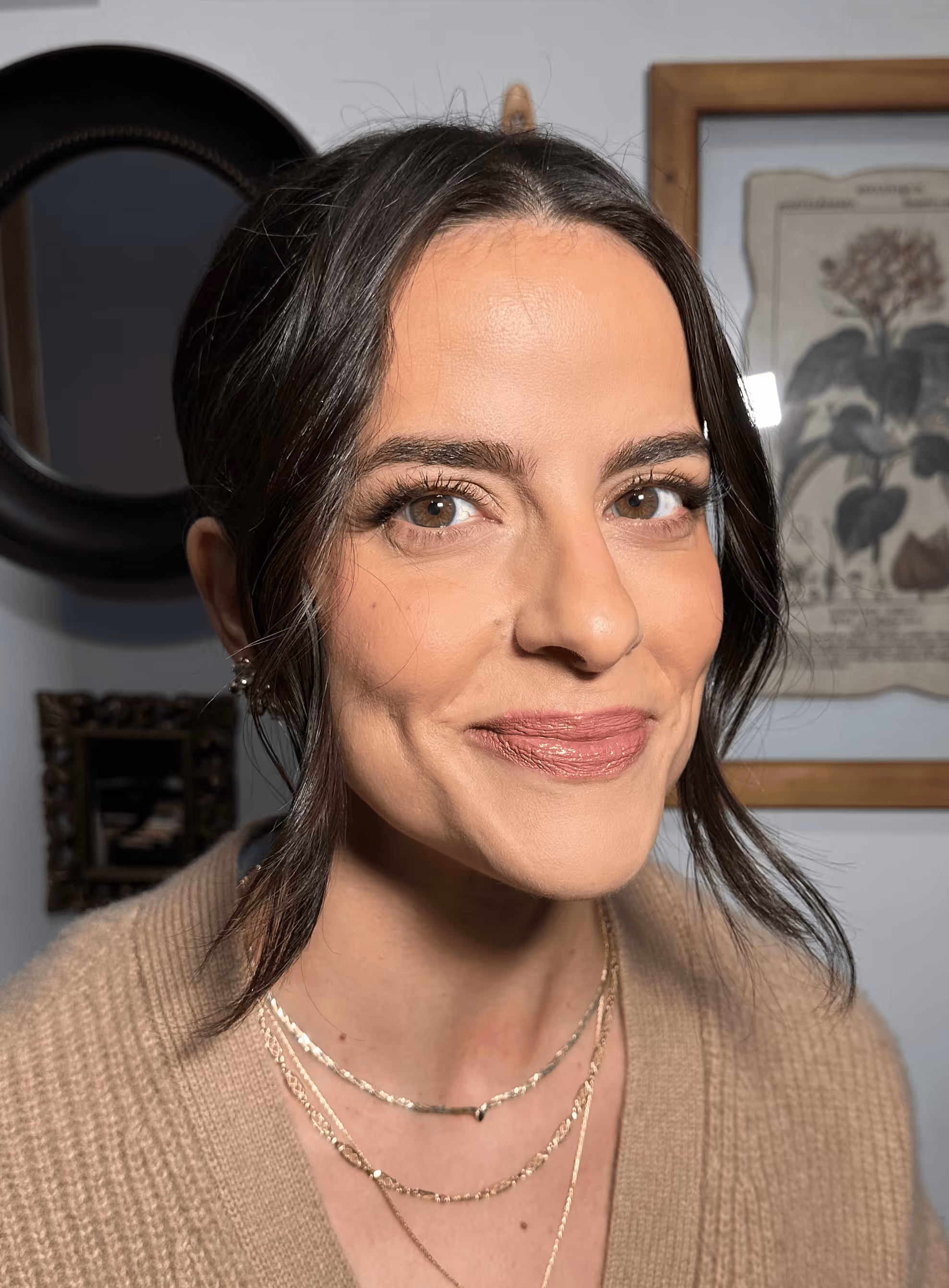 Smiling woman with brown hair in a beige sweater, wearing layered gold necklaces against a background with framed artwork.