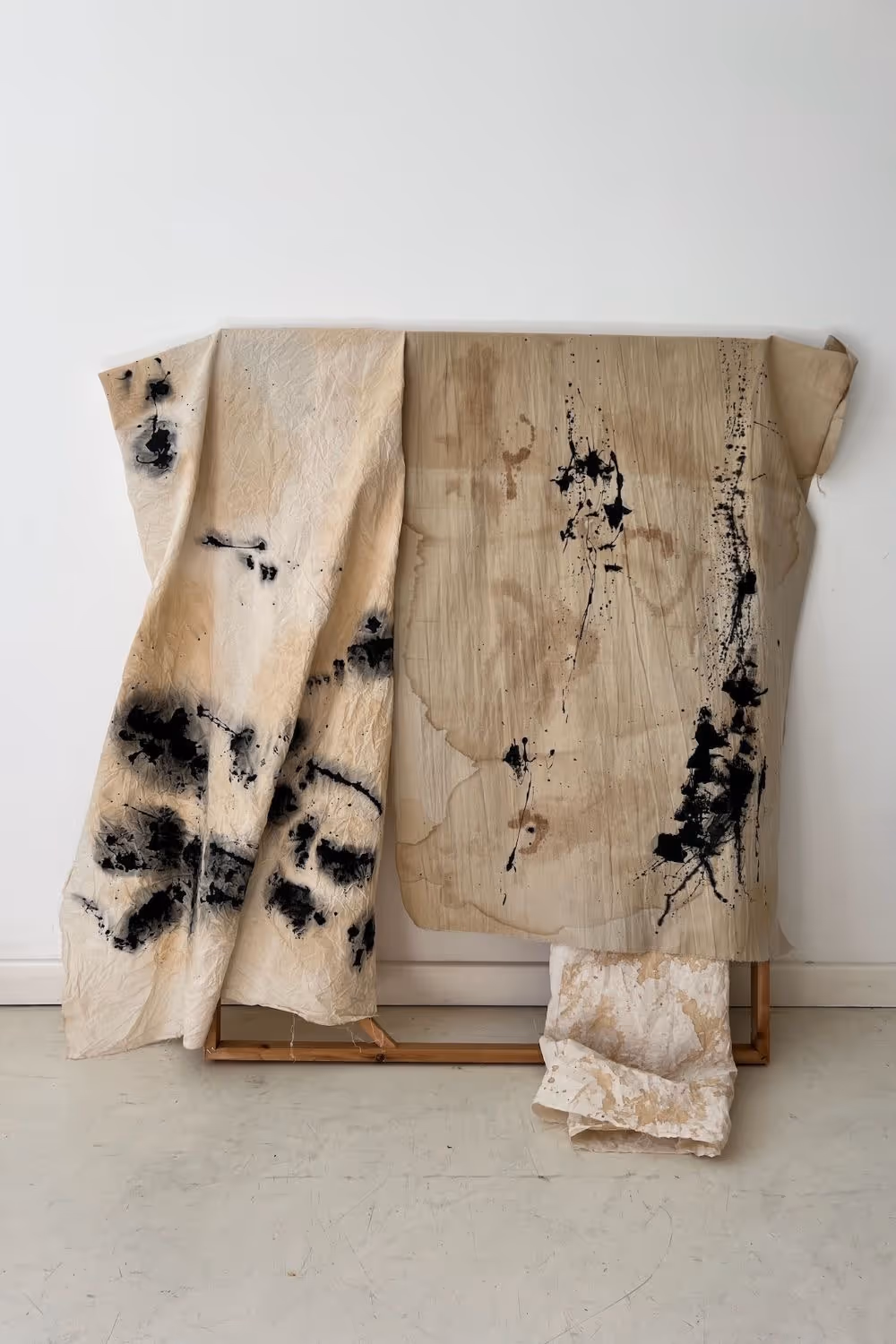 Three pieces of wrinkled fabric with black and brown ink stains draped over a wooden frame against a white wall.