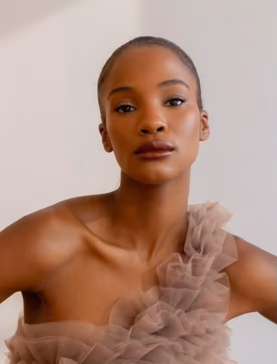 Portrait of a woman with smooth skin and dark lipstick wearing a ruffled tulle dress against a plain background.