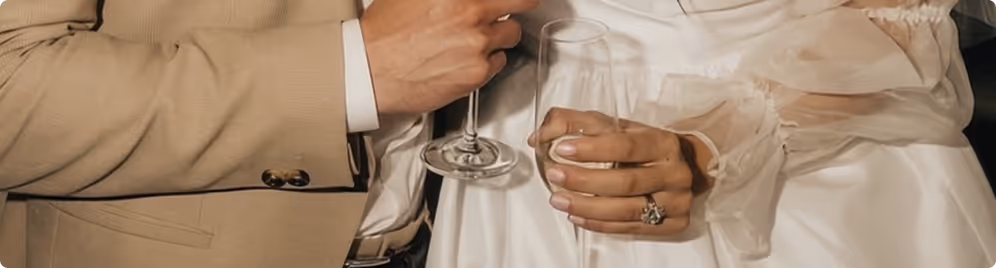 Close-up of a couple's hands holding champagne glasses, with the woman wearing a white sheer long-sleeve dress and a large engagement ring.