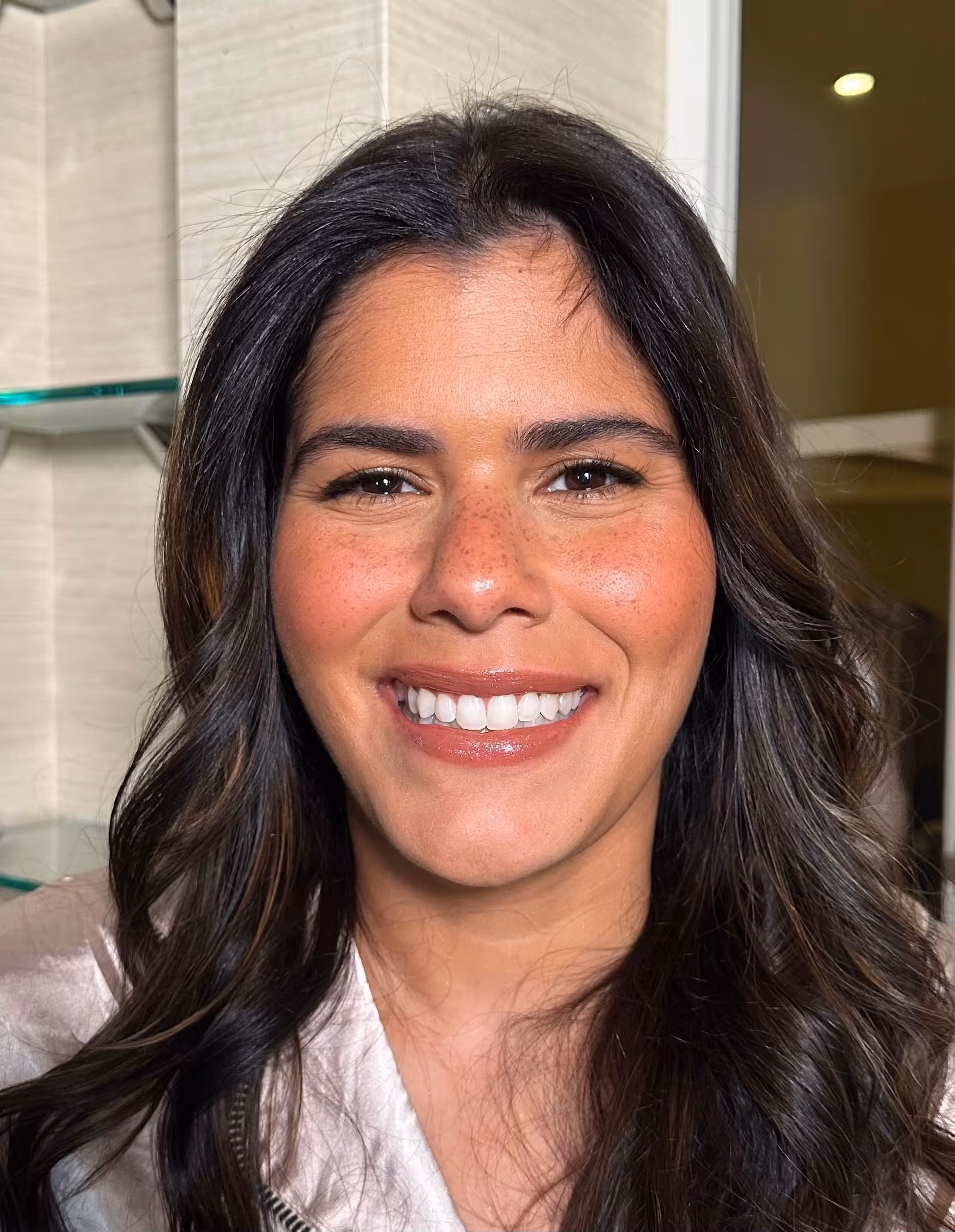 Smiling woman with long dark wavy hair, light freckles, and glossy lips in an indoor setting.
