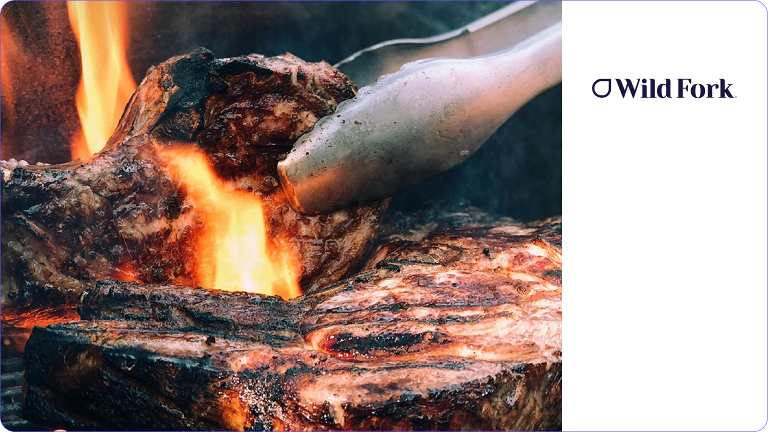Close-up of steak being grilled with tongs over open flames.