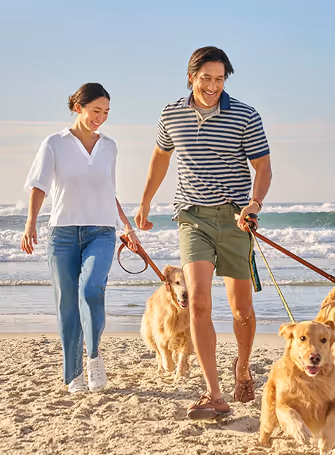 Smiling couple walking two golden retrievers on leashes along a sandy beach with ocean waves in the background.