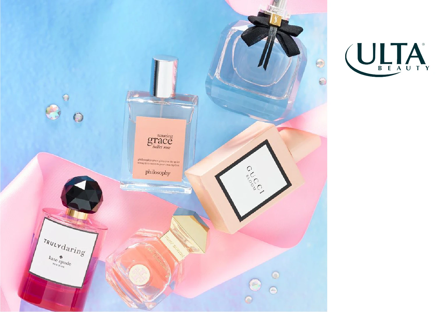 Flat lay of five luxury perfume bottles including Philosophy Amazing Grace, Gucci Bloom, Kate Spade Truly Daring, and two others on a blue and pink fabric background with small decorative gems.