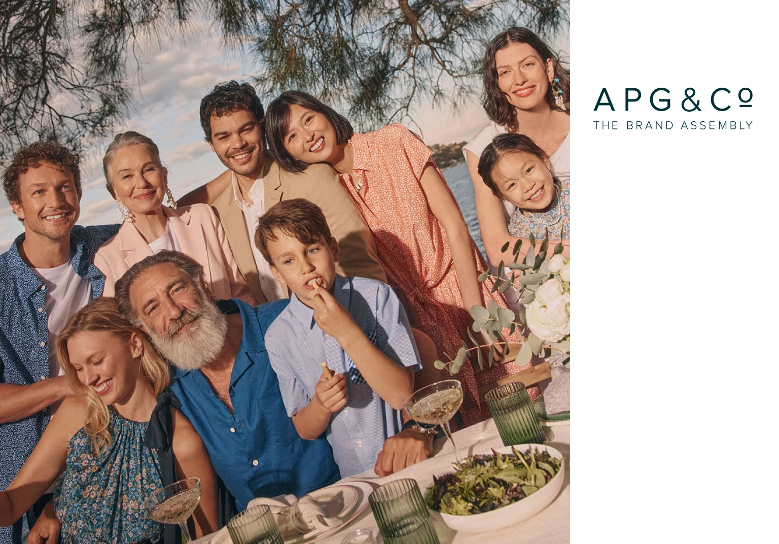Smiling diverse group of nine people, including adults and children, gathered outdoors around a dining table with food and drinks.