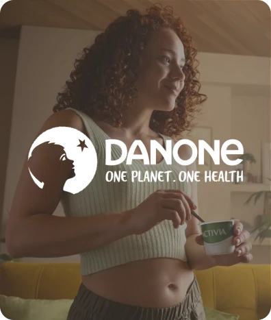 Woman with curly hair eating a cup of Activia yogurt in a cozy living room, with Danone logo and slogan 'One Planet. One Health' overlaid.