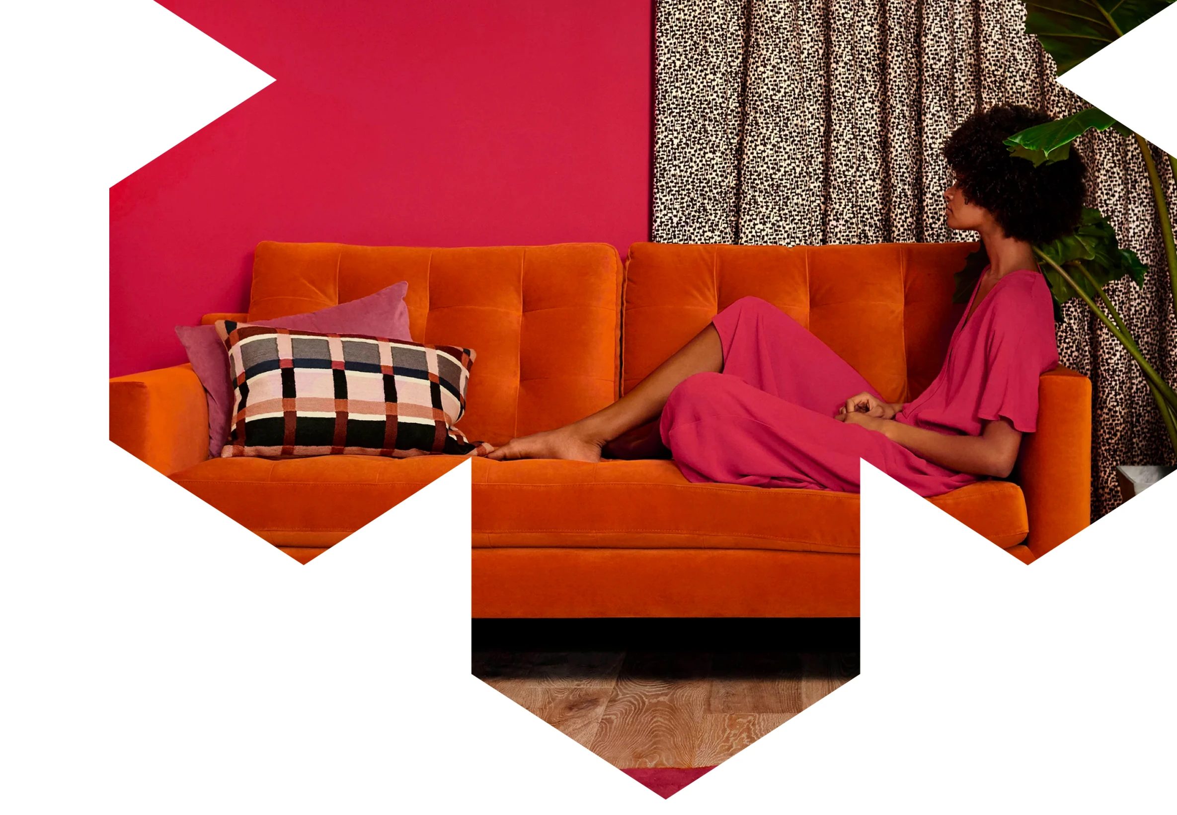 Woman with curly hair in a pink dress reclining on an orange sofa with patterned cushions, beside a leafy plant and leopard print curtains.