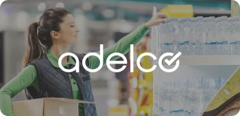 Woman in a green shirt and black vest placing a box on a shelf in a warehouse or store setting with water bottles on the shelf.