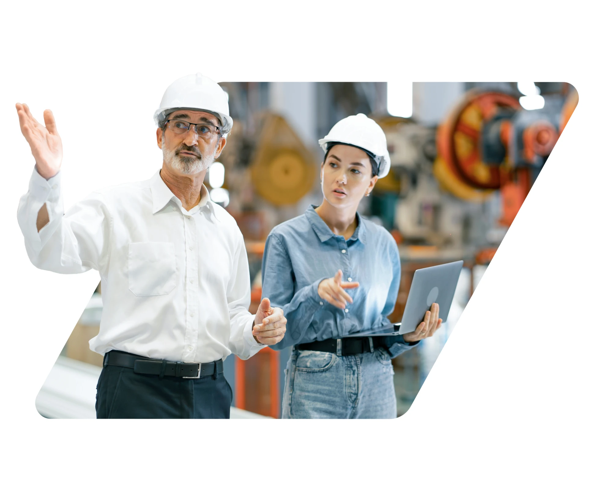 Two engineers wearing white safety helmets discussing while holding a laptop in an industrial factory setting.