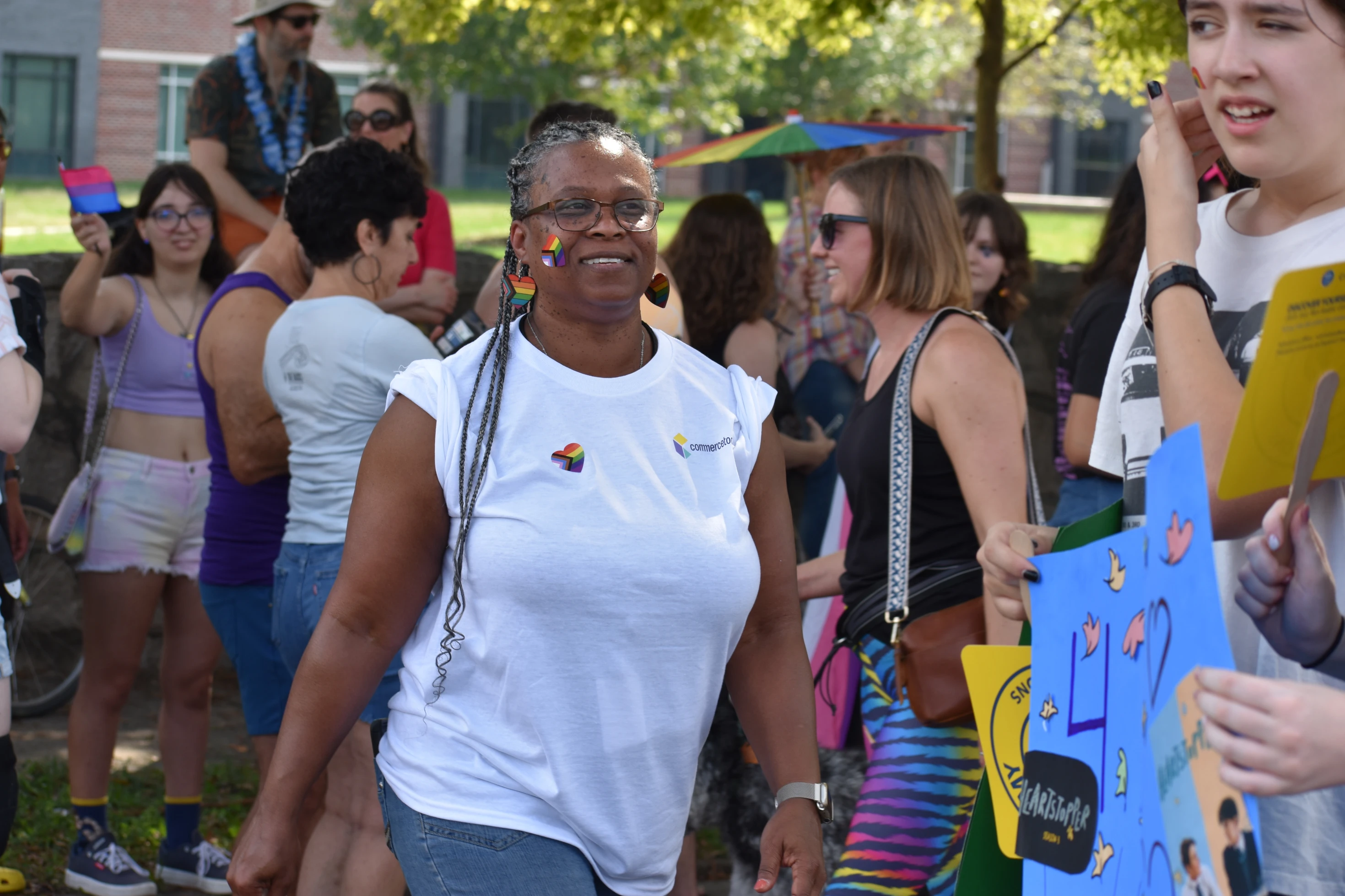 A smiling woman with rainbow pride stickers on her face and earrings, wearing a white shirt and walking in an outdoor gathering with diverse people holding colorful signs.