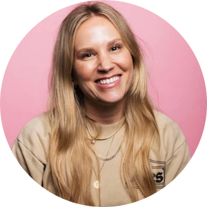 Smiling woman with long blonde hair wearing a beige top and layered necklaces against a pink background.