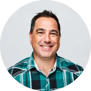 Smiling man with short dark hair wearing a green and black plaid shirt against a light gray background.