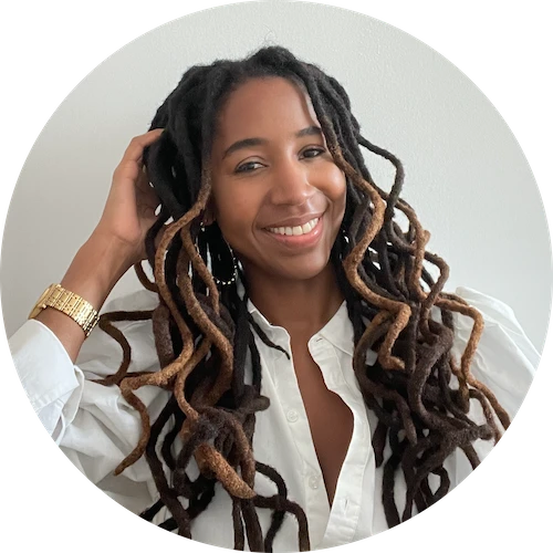 Smiling woman with long brown and black dreadlocks wearing a white shirt and gold watch.