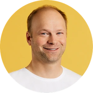 Smiling man with light skin, short light brown hair, and slight beard stubble, wearing a white shirt against a yellow background.