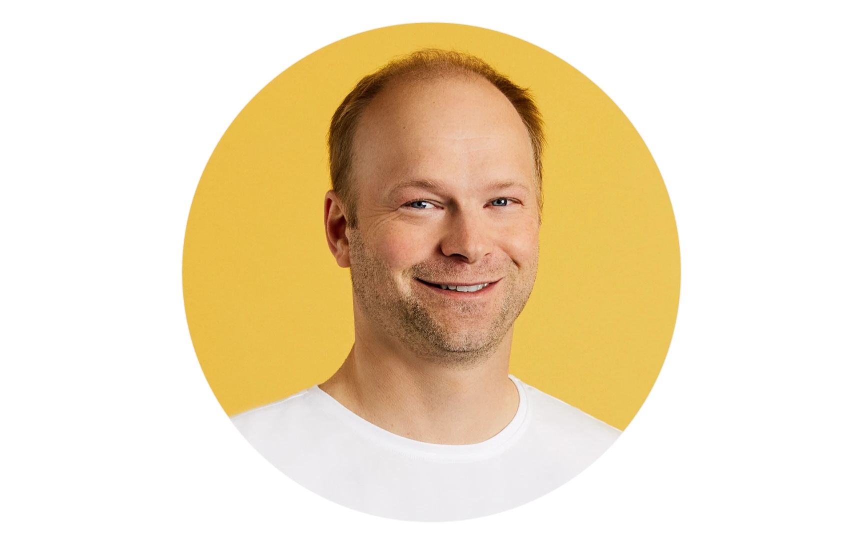 Smiling man with light skin, short light brown hair, and blue eyes wearing a white shirt against a yellow background.