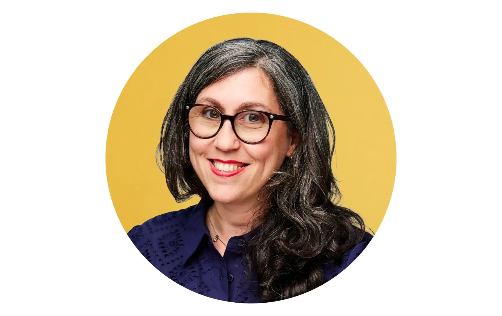 Smiling woman with long gray-streaked hair, wearing glasses and a dark blue shirt against a yellow background.