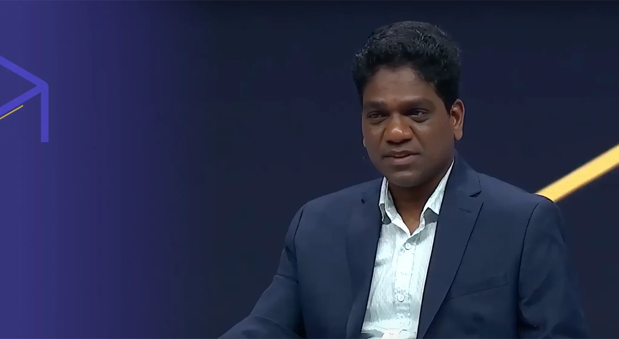Man with dark curly hair wearing a navy blue blazer and light patterned shirt against a dark purple background with geometric lines.