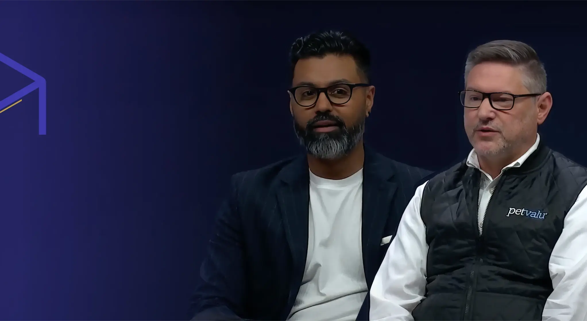 Two men wearing glasses seated against a dark blue background, one in a black blazer and white shirt, the other in a black vest with a petvalu logo over a white shirt.