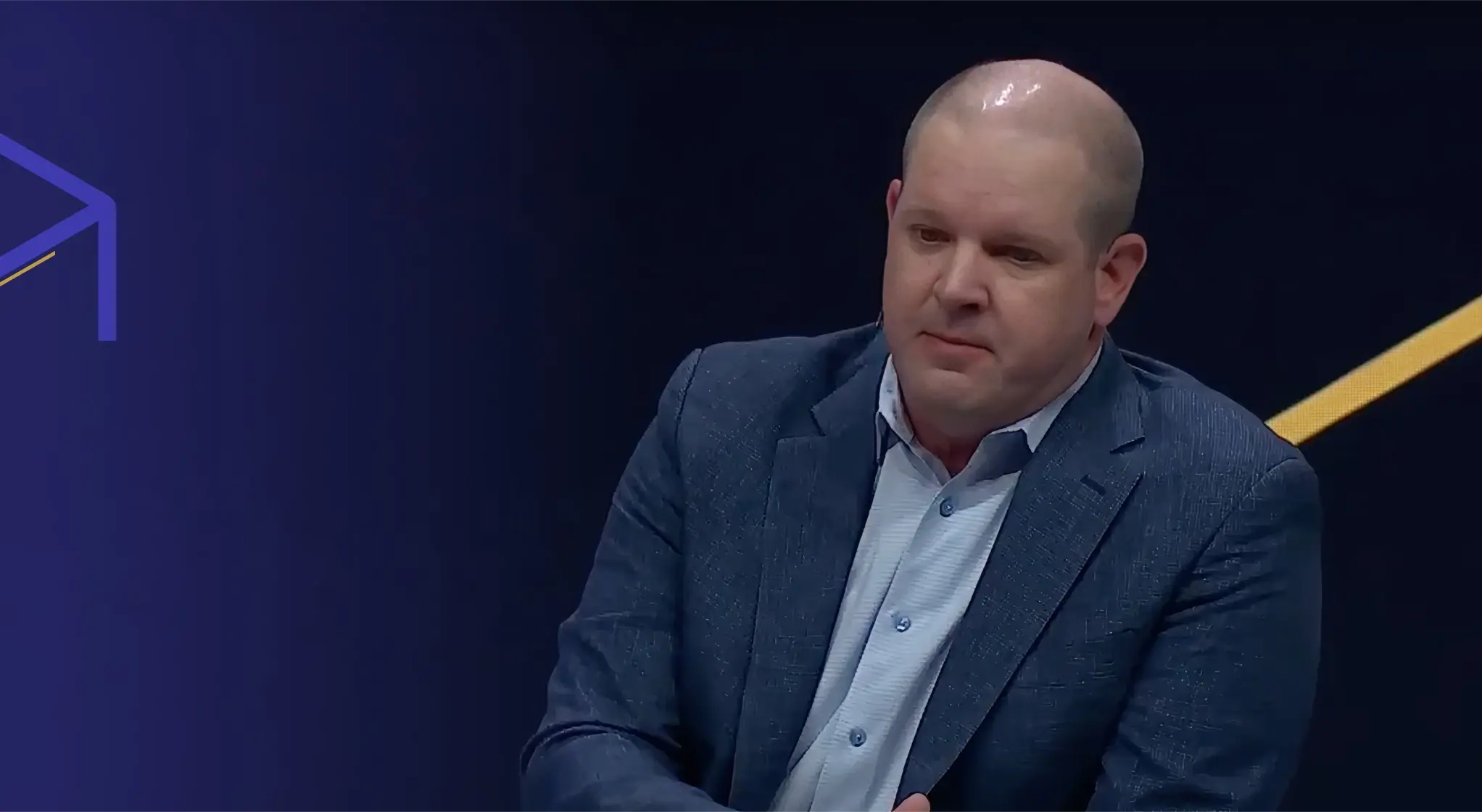 Man in a blue blazer and light blue shirt against a dark background with purple and yellow geometric lines.