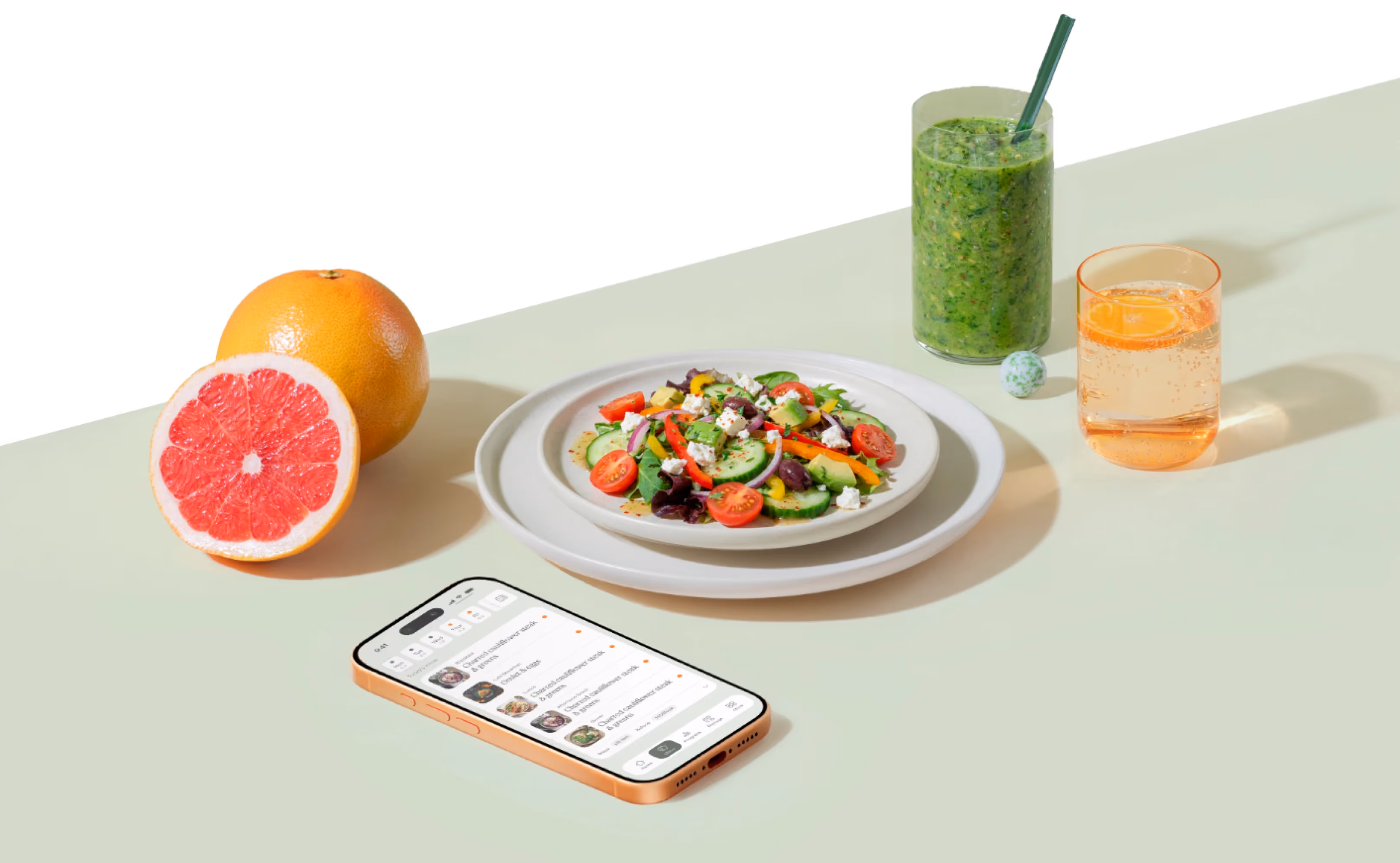 Talerz z sałatką z pomidorów koktajlowych, ogórka, sera feta i oliwek, obok pokrojony grejpfrut, zielony koktajl, szklanka z napojem i smartfon z aplikacją BeDiet Catering.